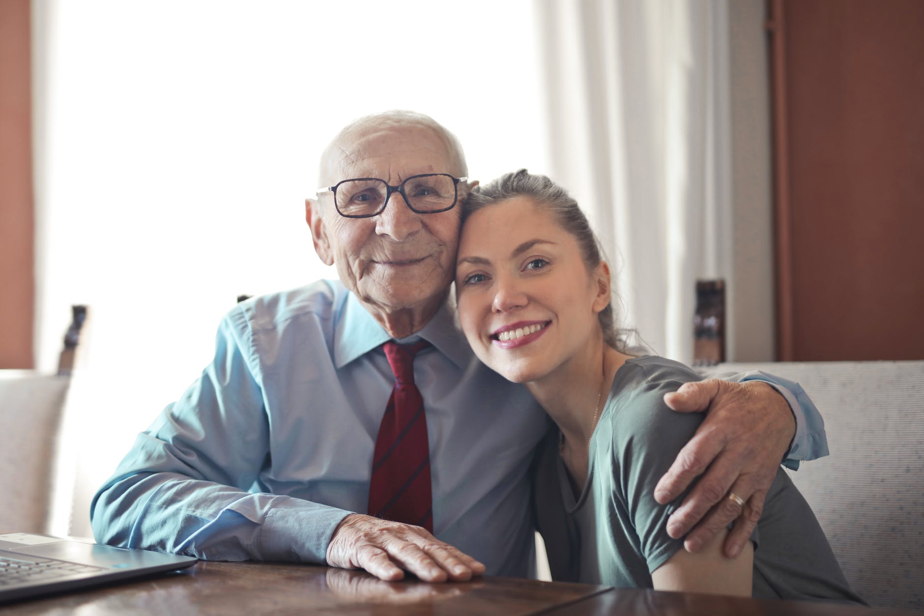 positive senior man in formal wear and eyeglasses hugging with young lady while sitting at table